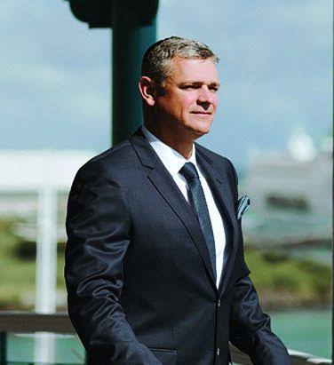 Arnaud Lagesse standing in a dark suit with the Mauritian coastline behind him